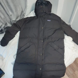 Svart lång dunjacka från Patagonia - Säljer en svart lång dunjacka från Patagonia med huva och tryckknappar framtill. Jackan är perfekt för kalla dagar och har en klassisk, stilren design med diskret logga på bröstet.