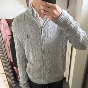 Grå stickad kofta från Polo Ralph Lauren - Säljer en grå stickad kofta från Polo Ralph Lauren med dragkedja och kabelstickat mönster. Tröjan har lång ärm, hög krage och den klassiska loggan broderad på bröstet. Perfekt för lager-på-lager-stil. Size S men något stor på mig så tycker den lutar mer mot Stor M/Liten L. Fint skick! För frågor kom pm! / Neo💫
