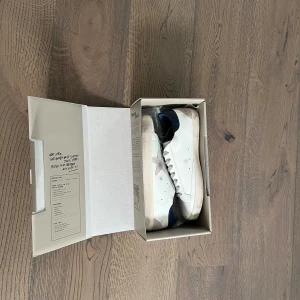 Golden Goose sneakers med blå detaljer storlek 43 - Säljer ett par vita Golden Goose sneakers med blå häl och grå mockadetaljer baktill. Skorna har klassisk snörning och en platt sula. Perfekta för dig som gillar stilrena och trendiga sneakers med en unik touch. Pris kan diskuteras vid snabbaffär. 