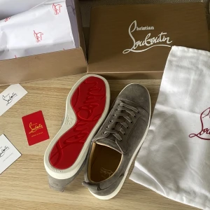 Gråa sneakers från Christian Louboutin - Säljer ett par gråa sneakers från Christian Louboutin med klassisk röd sula och snörning. Skorna är i mocka och har en rund tå samt platt sula. Perfekta för dig som vill ha något stilrent men ändå lyxigt. Helt nya som aldrig är använda eller testade. Alla tillbehör medföljer 