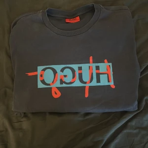 Mörkblå sweatshirt från Hugo Boss - Säljer en mörkblå sweatshirt från Hugo Boss med stort tryck i rött och blått framtill. Tröjan har rund halsringning och långa ärmar. Perfekt för en avslappnad och stilren look. Strl L passar även M