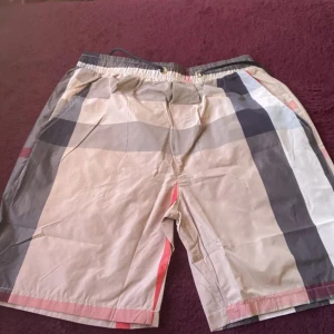 Beige shorts rutiga - Snygga beige shorts med stort rutigt mönster i svart, grått, vitt och rött. Shortsen har elastisk midja med snörning och en bakficka. Perfekta för sommaren och har en avslappnad passform.