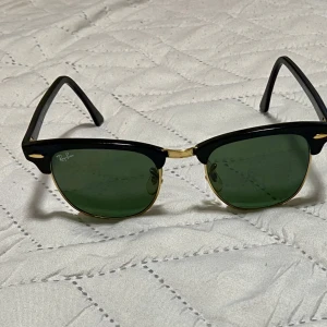 Ray-Ban Clubmaster solglasögon - Snygga Ray-Ban Clubmaster solglasögon med svart ovandel, gulddetaljer och gröna glas. Klassisk och tidlös design med metall och plast i bågarna. Perfekta för dig som vill ha en ikonisk look. Revan på fodral solglasögon knapp är tydlig,