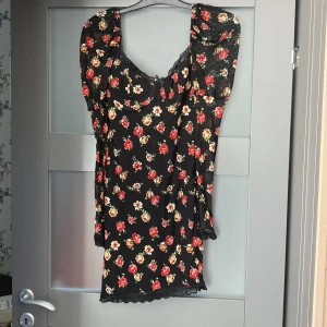 Svart blommig kortklänning från Kiss The Sky - Supersöt svart kortklänning från Kiss The Sky med röda och vita blommor. Klänningen har puffiga korta ärmar, spetsdetaljer vid halsringningen och nederkanten samt en figurnära passform. Perfekt för dig som gillar romantisk stil.