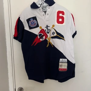 Flerfärgad piké t-shirt från Reason - Säljer en snygg piké t-shirt från Reason i vitt, marinblått och rött. T-shirten har korta ärmar, krage och flera broderade detaljer samt tryck, bland annat flaggor och siffran 6. Perfekt för dig som gillar sportig stil.