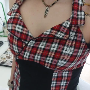 Rutig halternecktopp - Säljer en snygg röd, svart och vit rutig topp med halterneck. Toppen har svarta paneler i midjan med korsett design. Perfekt för dig som gillar en alternativ stil!