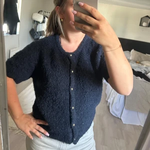 Mörkblå kofta med pärlknappar - Säljer en fluffig mörkblå kofta med korta ärmar och pärlknappar framtill. Perfekt att slänga på sig över ett linne eller t-shirt. Koftan har en mjuk och mysig känsla och passar till många olika outfits.