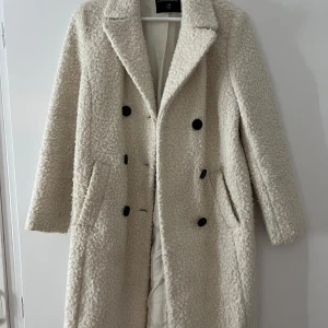 Vit/krämvit teddykappa från Dorothy Perkins - Säljer en lång, vit teddy kappa från Dorothy Perkins i storlek UK 6 (EU 34). Kappan har dubbelknäppning med svarta knappar, stora fickor och klassisk krage.