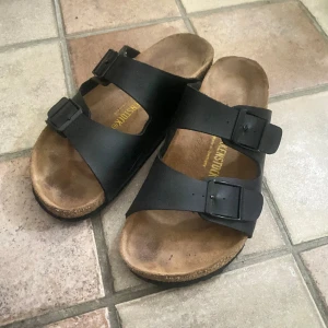 Svarta sandaler från Birkenstock - Klassiska svarta sandaler från Birkenstock med två breda remmar och justerbara spännen. Fotbädden är anatomiskt utformad och sulan är i kork för extra komfort. Perfekta för varma dagar och enkel att matcha med olika outfits.