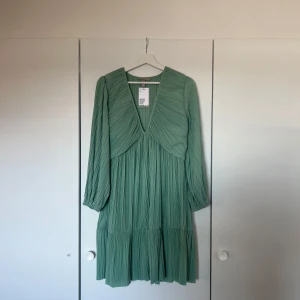 Grön plisserad klänning med v-ringning - Säljer en ljusgrön klänning med plisserat tyg och v-ringad hals. Klänningen har långa ärmar och en lös passform, perfekt för vår och sommar. Den har en luftig känsla och faller fint med volangdetalj nedtill. Den är helt ny och lappen finns fortfarande kvar, säljer den då den inte har kommit till användning🧊💕🫧
