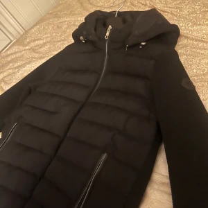 Svart pufferjacka med huva - Säljer en svart pufferjacka med huva och dragkedja framtill. Jackan har quiltade partier på framsidan och två fickor med dragkedja. Perfekt för kyligare dagar och enkel att matcha med olika outfits.