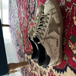 Vita sneakers från Golden Goose med grå stjärna - Säljer ett par vita sneakers från Golden Goose med grå stjärna på sidan och svarta detaljer vid hälen. Skorna har snörning och en platt sula. Märket 'SUPERSTAR' syns på innersulan och 'GGDB' på sidan.