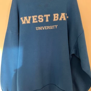 Blå sweatshirt med tryck 'WEST BAY UNIVERSITY' - Blå oversized sweatshirt från Weekday med vit text 'WEST BAY UNIVERSITY' på bröstet. Tröjan har rund halsringning och långa ärmar. Perfekt för en avslappnad stil och enkel att matcha med jeans eller leggings.