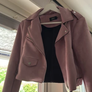 Lila bikerjacka från ONLY - Säljer en lila bikerjacka från ONLY i mockaimitation. Jackan har silvriga dragkedjor, knappar och klassiska detaljer som snedställd dragkedja och axelklaffar. Croppad modell med fickor framtill och dragkedjor vid ärmslut.