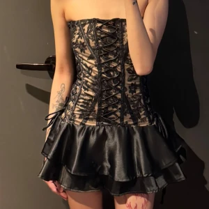 Svart axelbandslös kortklänning med snörning - Säljer en svart axelbandslös kortklänning med snörning framtill och blommiga spetsdetaljer. Kjolen är i glansigt material med volanger och omlottkänsla. Perfekt för dig som vill sticka ut med en edgy och snygg look.