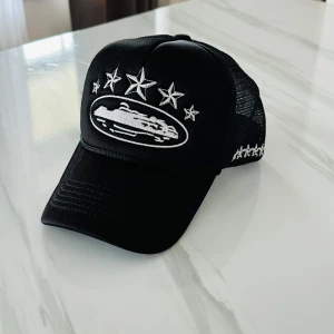 Svart Cortez keps  - Svart keps med vit broderad design framtill som föreställer ett fordon och flera stjärnor. Kepsen har mesh på sidorna och fler broderade stjärnor på sidan. Justerbar passform baktill.