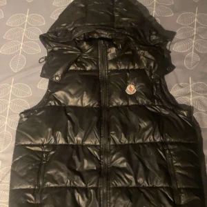 Svart dunväst från Moncler - Säljer en svart dunväst från Moncler med huva och dragkedja framtill. Västen har Moncler-logga på bröstet och två fickor med dragkedja. Perfekt för lager-på-lager och snygg till många outfits. Pris kan diskuteras kom med förslag 