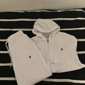 Vita byxor och hoodie från Ralph Lauren - Säljer vita byxor och hoodie från Ralph Lauren med dragkedja och huva. Tröjan har långa ärmar och den ikoniska lilla broderade loggan på bröstet. Perfekt för en avslappnad stil.