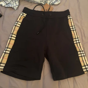 Svarta shorts med rutiga sidopaneler från Burberry - Snygga svarta shorts från Burberry med klassiska beige-rutiga sidopaneler. Shortsen har elastisk midja med snörning och är tillverkade i bomull för en bekväm känsla. Perfekta för en stilren och avslappnad look.