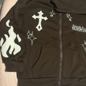 Svart hoodie med broderade detaljer - Svart hoodie med dragkedja och broderade vita motiv som kors, eldflammor och stjärnor. Stora fickor framtill och huva. Snygg streetwear-stil med unika detaljer på både ärmar och bröst.