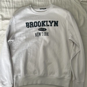 Vit sweatshirt med Brooklyn-tryck från Reaction - Säljer en vit sweatshirt från Reaction i storlek M med svart texttryck 'Brooklyn State of New York' på bröstet. Tröjan har rund hals, långa ärmar och ribbade muddar vid ärmslut och nederkant. Perfekt för en avslappnad stil.