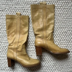 Beiga skinnstövlar med klack från Bronx - Säljer ett par beiga stövlar i skinn från Bronx med  klack på 7,5cm. Stövlarna har en klassisk design med dragflik upptill och markerade sömmar längs skaftet. Perfekta för dig som vill ha ett par stilrena boots med lite höjd. Mjukt och väldigt bekvämt läder. Har varit älskade och har lite skvätt från att ha gått. 