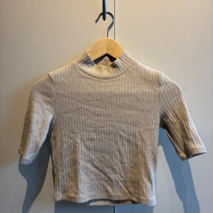 Beige ribbad topp med halv polokrage - Säljer en beige ribbad topp med halv polokrage och korta ärmar. Toppen har en tight passform och är perfekt för att styla med jeans eller kjol. Enkel och stilren design som passar till många tillfällen.