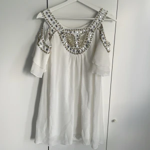 Vit klänning med pärldetaljer - Vit med öppna axlar klänning och glittrande pärlor och nitar längs urringningen och axelpartiet. Klänningen har volangärmar och ett luftigt, lätt tyg som ger en romantisk känsla. Perfekt för speciella tillfällen. Liten fläck vid bröstet på höger sida.