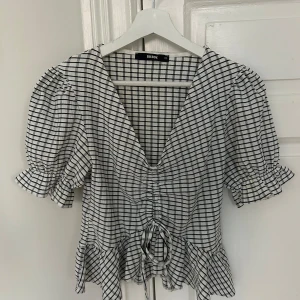 Rutig blus från Bik Bok - Säljer en söt rutig blus från Bik Bok i svart och vit. Blusen har korta puffärmar och en v-ringad design med knytning framtill.  Använd 1 gång. Priset kan diskuteras vid snabb affär😊