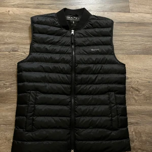 Svart dunväst från GANT - Svart GANT väst – storlek S Säljer denna stilrena och bekväma GANT-väst i storlek S. Den är lätt vadderad och perfekt som ett extra lager under vår och höst. Västen har två fickor med dragkedja och en mjuk ribbad krage för extra komfort. I mycket fint skick, då jag bara har testat den. Nypris 2200kr