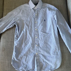 Blå skjorta från Polo Ralph Lauren - En klassisk blå skjorta från Polo Ralph Lauren med diskret rutmönster och broderad logotyp på bröstet. Skjortan har långa ärmar och knappar framtill.                       Inga konstigheter eller defekter 