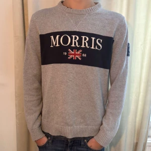 Grå tröja från Morris - Säljer en stilren grå tröja från Morris med ett broderat märke och brittisk flagga på framsidan. Tröjan har långa ärmar och ribbade muddar vid ärmslut och nederkant. Perfekt för en avslappnad stil.