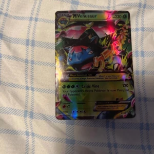 Pokémonkort M Venusaur EX - Säljer ett Pokémonkort med M Venusaur EX. Kortet har en färgglad design med M Venusaur i fokus och texten 'Crisis Vine' som attack. Perfekt för samlare eller Pokémon-entusiaster!