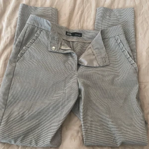 Rutig byxa från Zara - Snygga rutiga byxor från Zara i en klassisk grå nyans. Byxorna har en elegant volangdetalj vid fickorna och en smidig dragkedja framtill. Perfekta för en stilren look.