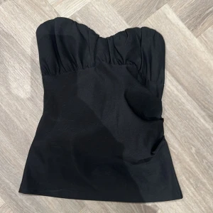 Svart axelbandslös topp - Snygg svart axelbandslös topp med rynkad detalj upptill. Perfekt för en stilren och elegant look. Passar bra till både jeans och kjol. Helt slutsålda, helt oanvänd