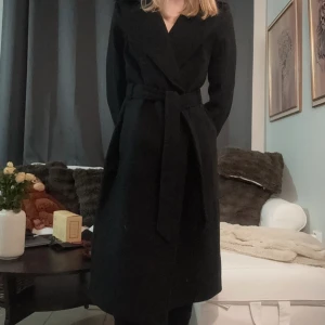 Svart kappa med bälte - Elegant svart kappa med bälte och omlott-design. Perfekt för kyligare dagar med sin långa ärm och klassiska stil. Passar bra till både vardag och mer uppklädda tillfällen.