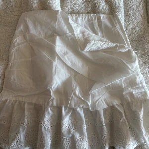 Vit kjol med broderade detaljer - Charmig vit kjol med vackra broderade blommönster och volangdetaljer. Perfekt för en somrig look. Kjolen har en omlott-design som ger en fin passform.