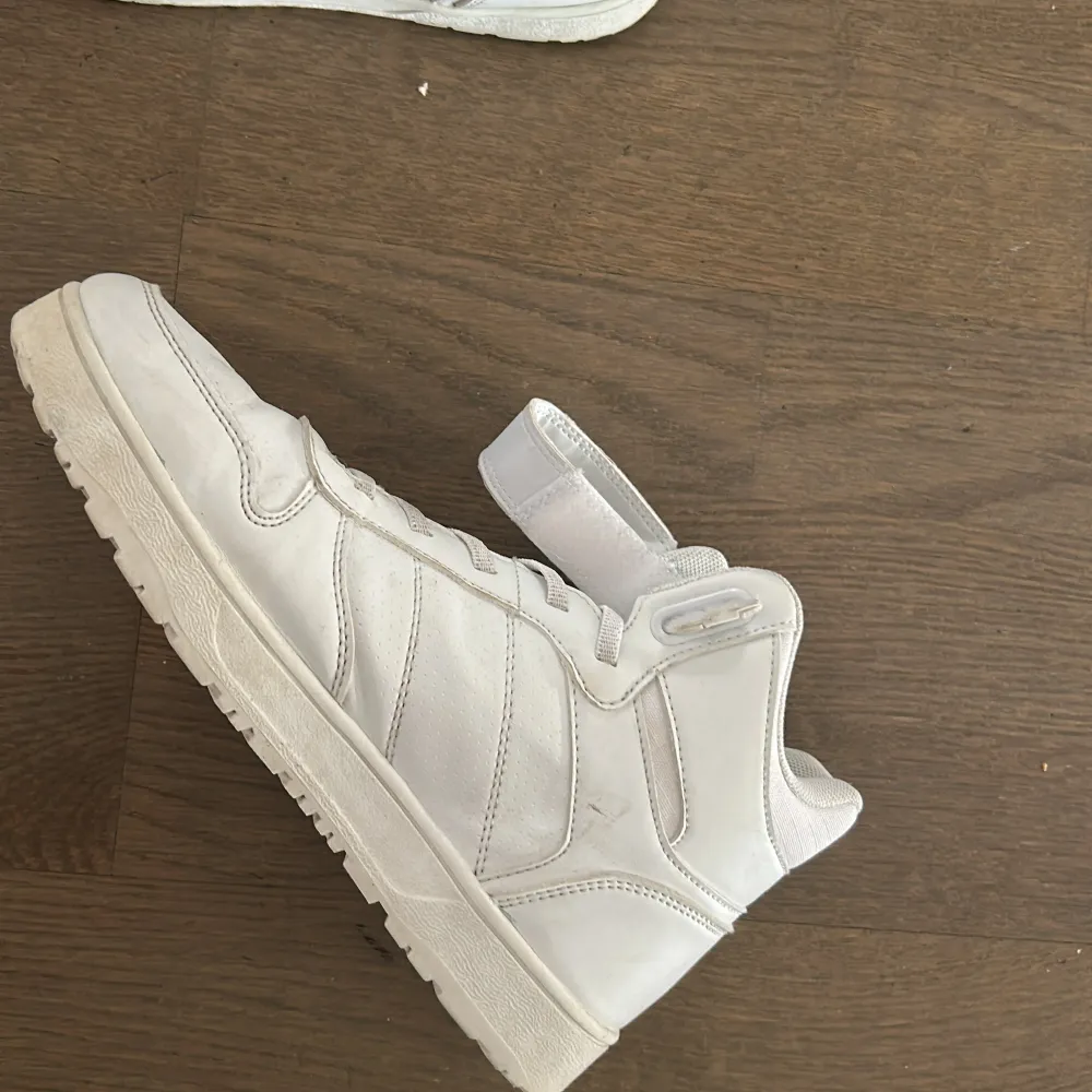 Snygga vita sneakers med hög modell och snörning. Skorna har även ett praktiskt kardborreband runt ankeln för extra stöd. Perfekta för en sportig look.. Kengät.