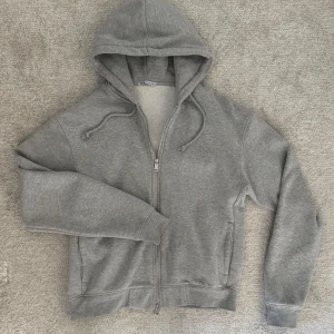 Grå hoodie från Zara - Säljer en stilren grå hoodie från Zara med dragkedja och justerbar huva. Perfekt för en avslappnad look. Den är tillverkad i mjukt material och har långa ärmar.