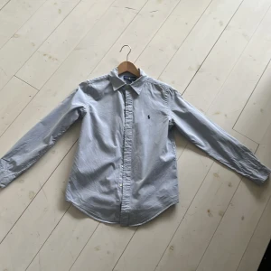 Ralph Lauren Skjorta - Säljer en stilren ljusblå skjorta från Ralph Lauren med klassisk krage och broderad logotyp på bröstet. Skjortan har långa ärmar och knappar framtill. Perfekt för en elegant look.