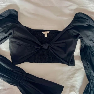 Svart kortärmad topp från H&M - Säljer en svart kortärmad topp från H&M med puffiga ärmar och smockdetaljer. Toppen har en knytning framtill och är perfekt för en trendig look.