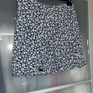 Blommig kortkjol - Söt kortkjol med blommigt mönster i blått och vitt på en svart bakgrund. Kjolen har en lätt utställd form och är perfekt för en somrig look. Den har en bekväm passform och är fodrad för extra komfort.