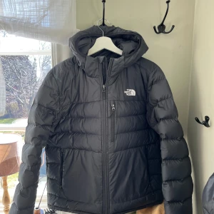 Svart dunjacka från The North Face - Säljer en svart dunjacka från The North Face. Jackan har en huva och dragkedja framtill. Perfekt för kalla dagar med sin quiltade design som håller värmen inne.
