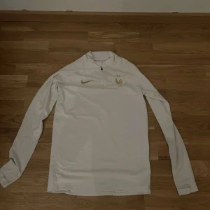 Frankrike tröja - Säljer en stilren vit långärmad tröja från Nike med dragkedja vid halsen och ett guldigt emblem på bröstet. Perfekt för sportiga aktiviteter eller som en snygg vardagströja. 🏃‍♂️