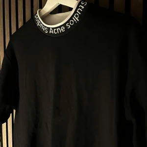 Svart t-shirt från Acne Studios - En clean svart t-shirt från Acne Studios med deras logga i vitt runt halsringningen. Perfekt för en stilren look. Tröjan har en klassisk passform och är tillverkad i ett mjukt material. 
