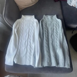 Vit och grå stickad polotröja - Två mysiga stickade polotröjor i vit och grå färg. Båda har ett klassiskt flätmönster och långa ärmar, perfekta för kyligare dagar. Polokragen ger extra värme och stil. Den gråa är xs och den vita S.  1 för 150kr. Båda för 250kr
