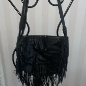 Svart axelväska med fransar - Snygg svart axelväska i skinn med coola fransdetaljer. Väskan har en dragkedja upptill och en praktisk snörning framtill. Perfekt för att ge din outfit en edgy touch.