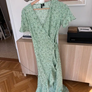 Grön blommig omlottklänning från Vero Moda - Säljer en söt grön omlottklänning från Vero Moda med blommigt mönster. Klänningen har korta ärmar och knytning i midjan för en smickrande passform. Perfekt för vår och sommar!