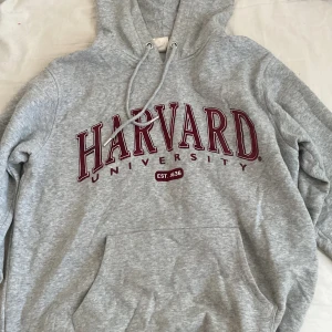 Grå Harvard hoodie - Säljer en klassisk grå hoodie med Harvard University-tryck i vinrött. Hoodien har en stor ficka framtill och justerbar huva med dragsko. Perfekt för en avslappnad stil eller som ett statement-plagg.