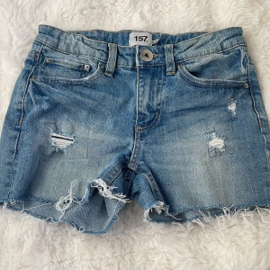 Blå jeansshorts med slitningar från Lager 157 - Snygga blå jeansshorts från Lager 157 med slitna detaljer och fransig kant.  De har en knapp och dragkedja framtill samt klassiska fem fickor. Dem är i storlek 150. Midja rakt över: 33cm.